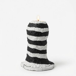 May include: A cylindrical candle with a textured, uneven surface featuring horizontal black and white stripes. The candle has a wide, flat base and a single lit wick at the top, emitting a small, warm flame against a plain white background.