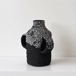 May include: A black vase with a textured, matte surface and two side handles. The upper portion of the vase and the handles feature a dense pattern of small, white, hand-painted vertical dashes that contrast against the solid black base.