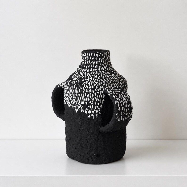 May include: A black vase with a textured, matte surface and two side handles. The upper portion of the vase and the handles feature a dense pattern of small, white, hand-painted vertical dashes that contrast against the solid black base.
