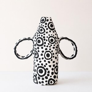 May include: A white ceramic vase with a textured surface featuring a hand-painted black pattern of various sized circles and dots. The vessel has a tall, cylindrical body that tapers towards the top and two large, circular handles on each side.