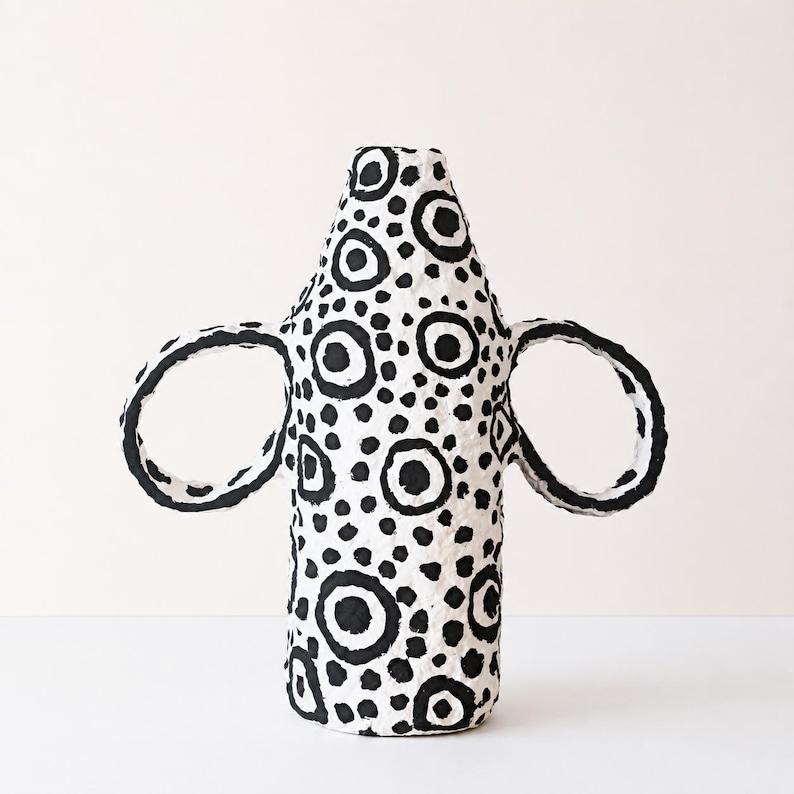 May include: A white ceramic vase with a textured surface featuring a hand-painted black pattern of various sized circles and dots. The vessel has a tall, cylindrical body that tapers towards the top and two large, circular handles on each side.