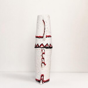 May include: A tall, cylindrical white vase with a rough, textured surface. It features hand-painted red and black geometric patterns, including horizontal bands and triangular motifs, along with small protruding decorative elements on the side.
