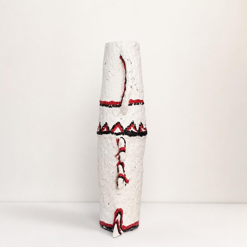 May include: A tall, cylindrical white vase with a rough, textured surface. It features hand-painted red and black geometric patterns, including horizontal bands and triangular motifs, along with small protruding decorative elements on the side.
