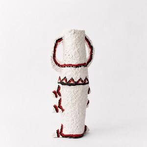 May include: A tall, textured white vase with a rough, paper-like surface. It features two large handles and decorative geometric patterns painted in bold black and red lines. The design includes triangular motifs and solid borders around the base.