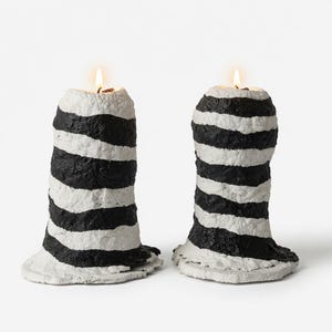 May include: Two cylindrical candles with a rough, textured surface featuring horizontal black and white stripes. Each candle has a lit wick at the top, emitting a warm glow. The base of each candle is slightly flared and uneven, showing a hand-molded appearance.
