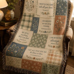 May include: A woven throw blanket featuring a patchwork design of floral patterns, plaid squares, and religious text including Trust in the Lord, Be Still, His Mercies, and Let All That You Do Be Done in Love, finished with a fringed edge.