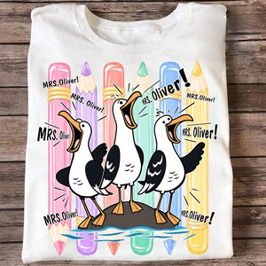 May include: A white t-shirt featuring a graphic of three seagulls standing on a rock, each with an open beak. Behind the birds are five vertical colored pencils in pink, purple, coral, teal, and yellow. The text MRS. Oliver! appears repeatedly.