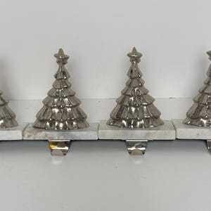 May include: A set of four stocking holders, each featuring a metallic silver Christmas tree figurine mounted on a rectangular white marble base with a polished silver metal hook extending from the underside for hanging holiday decorations.