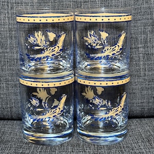 May include: A set of four clear glass tumblers featuring a repeating blue and cream-coloured bird and floral motif. Each glass has a horizontal band near the rim with a dotted pattern and a solid blue line, complemented by a matching blue base rim.