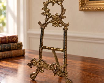 May include: A gold-toned metal display easel featuring an ornate, scrollwork top crest and a horizontal support bar. The structure has two front-facing feet with floral accents and a rear-hinged leg for stability, finished with a weathered texture.