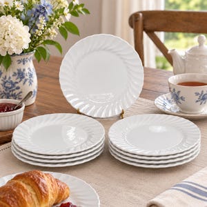 May include: A set of white ceramic plates featuring a distinct spiral or swirl pattern along the rim. The collection includes one upright plate displayed on a stand and two stacks of smaller plates, all characterized by a glossy finish and smooth texture.