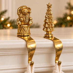 May include: Two gold-toned metal stocking holders with a polished finish. One holder features a bear wearing a hat and scarf sitting on a drum, while the other is shaped like a Christmas tree topped with a star. Both have curved hooks.