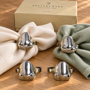 May include: A set of four silver-toned metal frog napkin rings, each featuring a stylized frog head design. Two rings are placed on cream-colored fabric napkins, and two are on sage green napkins. A tan box labeled Pottery Barn is behind them.