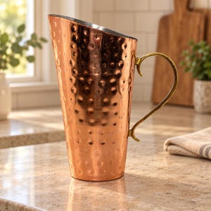 May include: A tall, tapered copper pitcher featuring a hammered texture across its exterior surface. The vessel includes a curved, metallic handle attached to the side and a wide, angled spout at the top for pouring liquids.