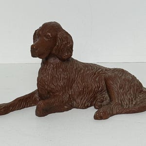 May include: A solid brown figurine of a dog in a lying position. The sculpture features detailed molded fur texture, long floppy ears, and a focused expression. The dog is depicted resting with its front legs extended forward and its tail tucked.