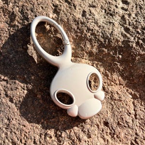 May include: A smooth, light beige carabiner keychain featuring a stylized, rounded skull-like shape with two circular eye cutouts and a prominent, curved top loop for attachment. The object has a glossy, solid finish and a uniform, neutral color.