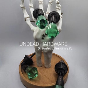 May include: Four green glass cabinet knobs with a dark bronze finish. The knobs are displayed on a skeletal hand and on a wooden base. The text "UNDEAD HARDWARE By Undead Furniture Co" is visible below the hand.