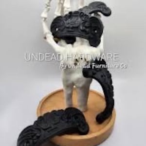 May include: Three black ornate metal drawer pulls with a decorative design. The pulls are displayed on a wooden surface.
