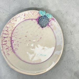 May include: A handmade ceramic plate with a blackberry design. The plate is off-white with a purple rim and accents. A sculpted blackberry with green leaves adorns the edge. The artist's signature, "J. Miller," is visible.