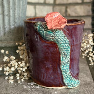 May include: A handmade ceramic mug with a deep burgundy glaze. A turquoise snake-like design wraps around the mug, topped with a coral-colored flower. The mug is a unique piece of art.