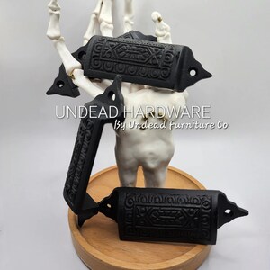 May include: Three black cast iron drawer pulls with an ornate design. The pulls are displayed on a wooden base and a skeletal hand.