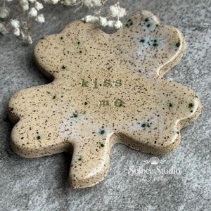 May include: A ceramic shamrock-shaped dish with a speckled beige finish and the words "kiss me" in green. The dish has a glossy surface and is decorated with small black and blue spots. The Solbee Studio logo is visible.