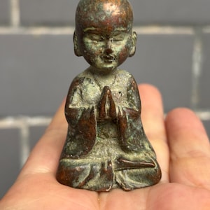 May include: A small, weathered bronze-toned figurine of a seated figure with a shaved head, depicted in a meditative pose with hands pressed together in prayer. The surface features a mottled patina with dark brown and oxidized green tones.