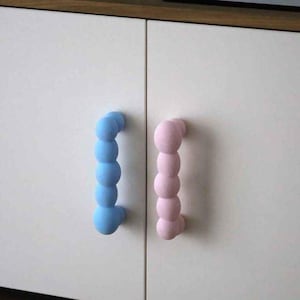 May include: Two vertical cabinet handles attached to white doors. One handle is light blue and the other is light pink. Both handles feature a series of five connected spheres in a linear arrangement, creating a textured, rounded, and tactile surface.