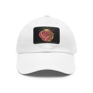 Puede incluir: Una gorra de béisbol blanca con un parche rectangular negro en la parte frontal. El parche muestra un gráfico de un corazón rojo estilizado entrelazado con una cruz de madera marrón, sobre un fondo circular amarillo pálido con líneas radiantes.