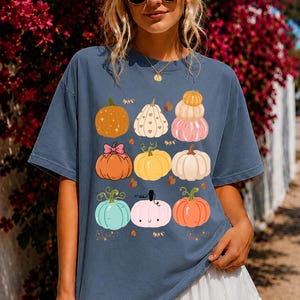 Puede incluir: Una camiseta de manga corta color azul pizarra con un estampado gráfico de nueve calabazas variadas en tonos naranja, amarillo, rosa, blanco y verde azulado. Las calabazas están dispuestas en cuadrícula, algunas decoradas con corazones, lazos y una cara sonriente.