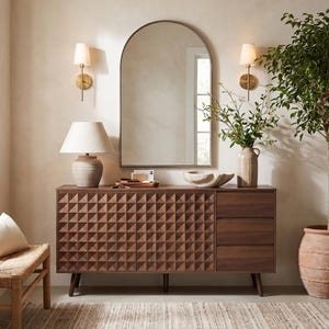 May include: A dark wood sideboard featuring a geometric pyramid-textured cabinet door and three smooth-fronted drawers. The piece stands on four tapered legs and is topped with a ceramic table lamp, a wooden tray, and a stone bowl.