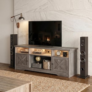 May include: A gray wooden TV stand with two side cabinets featuring X-shaped door panels, an open center shelf with integrated lighting, and a flat top surface supporting a large television screen, flanked by two tall black floor-standing speakers.