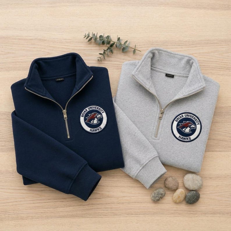 May include: A navy blue and a heather gray quarter-zip sweatshirt, each featuring a circular patch on the left chest with the text Briar University Hawks and a hawk head graphic. Both garments have long sleeves, ribbed cuffs, and a metal zipper.