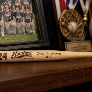 May include: A light-coloured wooden baseball bat featuring the engraved number 24, the word Bombers, the years 2025-2026, the name Owen Huntemer, and the number #24. The bat is positioned horizontally in front of a framed team photograph and a gold medal.