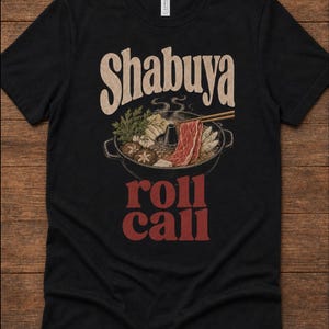 Puede incluir: Una camiseta negra de manga corta con un gráfico de una olla caliente con verduras, setas y rodajas de carne cruda. Sobre la olla, el texto "Shabuya" en color crema, y debajo de la olla, el texto "roll call" en una fuente roja negrita.