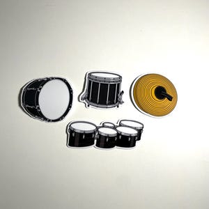 Puede incluir: Una colección de cuatro pegatinas de batería que incluyen un bombo negro, una caja negra, un platillo circular amarillo con detalles negros y un conjunto de cuatro toms negros con parches blancos, todos con bordes blancos.