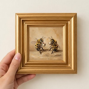 May include: A square painting in a gold-toned frame depicting two yellow and black striped bees sitting and holding small white cups. The artwork features a textured, neutral-toned background with visible brushstrokes, capturing the bees in a seated position as if drinking from the cups.
