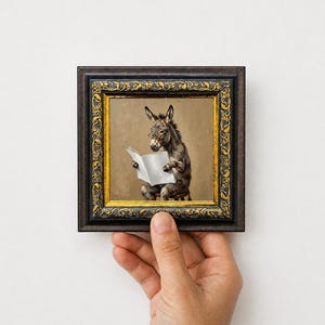May include: A square framed painting featuring a grey donkey wearing small spectacles while sitting on a stool and reading a white book. The frame is dark brown with an ornate gold inner border featuring embossed leaf and vine patterns.