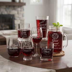 May include: A collection of six clear glass vessels filled with dark red liquid, each featuring white etched text and icons. The glassware includes a mason jar with a handle, a stemmed wine glass, and four tumblers, all labeled with Conference 2027.