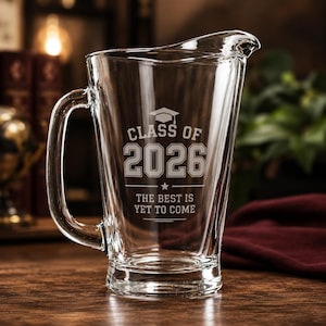May include: A clear glass pitcher featuring an etched design with a graduation cap icon, the text "CLASS OF 2026", and the phrase "THE BEST IS YET TO COME". The pitcher has a sturdy handle and a wide pouring spout, designed for serving beverages.