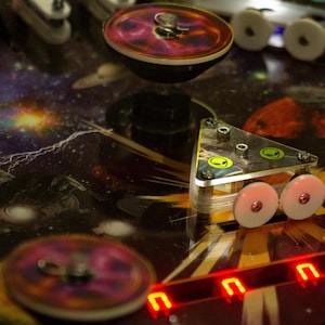 May include: A pinball machine playfield featuring a triangular bumper with two green alien head icons, white circular bumpers, and a space-themed graphic background with planets and stars. Bright red digital numbers are visible at the bottom right.