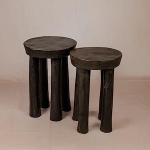 May include: Two dark charcoal-colored stools with circular tops and three thick, cylindrical legs each. The stools feature a matte, textured finish and are positioned side-by-side, with one stool slightly taller than the other in this set.