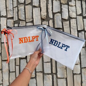 Könnte beinhalten: Zwei rechteckige, durchsichtige Reißverschlusstaschen aus Netzgewebe mit dem gestickten Schriftzug NDLPT. Eine Tasche zeigt orangen Text mit einem korallenfarbenen Band, die andere dunkelblauen Text mit einem blau-weiß karierten Band.
