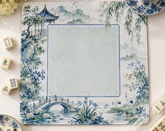 Op de afbeelding: Een vierkante mat met een aquarel-landschap met een pagode, een stenen brug, wilgentakken en kraanvogels. Het midden heeft een lichtblauw rechthoekig kader met een decoratieve rand. Mahjong-tegels en een theekopje liggen in de buurt.