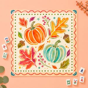 May include: A square decorative mat featuring a watercolor illustration of two pumpkins, one orange and one teal, surrounded by colorful autumn leaves, acorns, and berry sprigs. The border has a red scalloped edge and green dots.