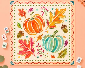 May include: A square decorative mat featuring a watercolor illustration of two pumpkins, one orange and one teal, surrounded by colorful autumn leaves, acorns, and berry sprigs. The border has a red scalloped edge and green dots.