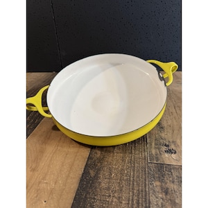 May include: A round, shallow cooking pan with a bright yellow exterior and a smooth white interior. The pan features two yellow handles on opposite sides and a thin black rim around the top edge, showing a small area of wear on the right side.