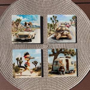 May include: A set of four square coasters featuring desert-themed illustrations. One shows a ram in a convertible, one a tortoise on a road, one a roadrunner wearing a bandana, and one a rabbit sitting outside a vintage camper trailer.