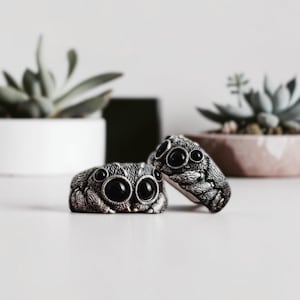 May include: Two silver-toned rings shaped like jumping spiders. Each ring features a detailed, textured metallic body with prominent, smooth, round black eyes set into the front. The design captures the distinct facial features of a spider.