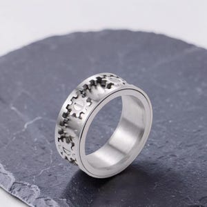 May include: A silver-coloured metal ring featuring a band design composed of interconnected gear shapes. The surface of the ring has a brushed metallic finish, and each individual gear element includes a central horizontal line detail.